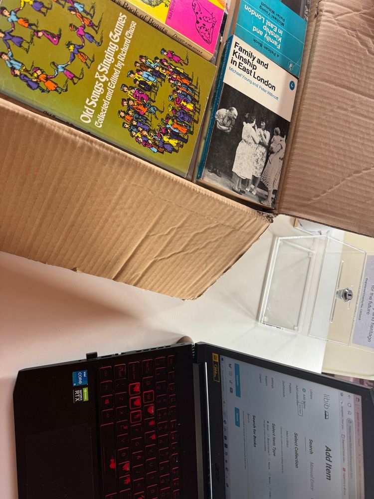 Photo of a laptop and part of a box of vintage books on a table. The vintage books are part of a large donation to The Folklore Library & Archive in Devon, and this box is full of books about childhood, language and children's lore. 