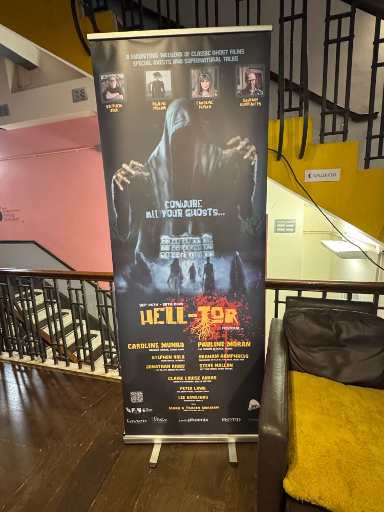 Roller banner advertising the 2025 Hell Tor Film Festival, featuring Caroline Munro, Pauline Moran, Stephen Volk, Graham Humphreys, Jonathan Rigby, Steve Nallon, Claire Louise Amias, Peter Laws, Lee Rawlings, Tracey Norman and Mark Norman. A hooded, faceless figure is reaching out towards the viewer. Below, a group of spectral figures walk through the ground fog towards a spooky-looking old house. Above them are the words 'Conjure all your ghosts'. 