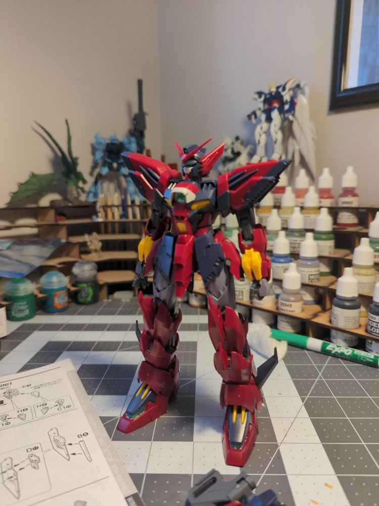 Gundam Epyon, a maroon and black Gundam with yellow accents that stands half finished on a craft table. 