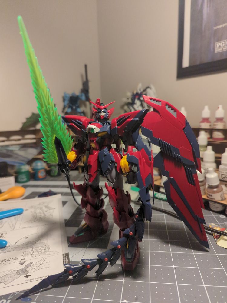 Gundam Epyon, a maroon and black Gundam with yellow accents stand completed with Large bat like wings, a neon green sword and shield with a chain whip. 