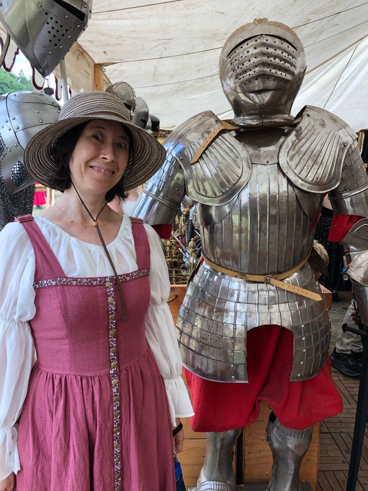 A photograph of author Kristina Fahl, 
with an excited expression on her face, wearing a pink/purple dress and a sun hat, standing next to a suit of armor. 