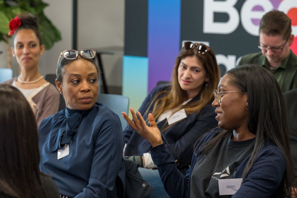 People seated indoors in discussion. One person in the foreground gestures while speaking. Faces are blurred. A colourful wall and partial sign reading 'Be' are visible in the background. Some attendees wear name tags and sunglasses on their heads.