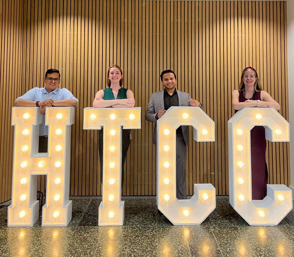 Four members of the Healthcare Technologies Capability Connector (HTCC) programme are standing behind large, illuminated letters that spell out "HTCC". The letters are white with light bulbs embedded in them. The background consists of vertical wooden slats. Each person is leaning on one of the letters