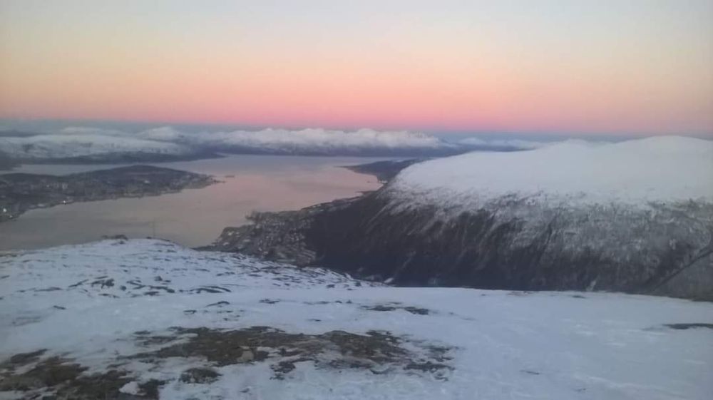 Image shows the view from the top of a mountain in Tromsø , Norway.  There is snow on top of all the mountains and a pink orange glow across the dusk sky. A wide river flows across the bottom of the valley. 