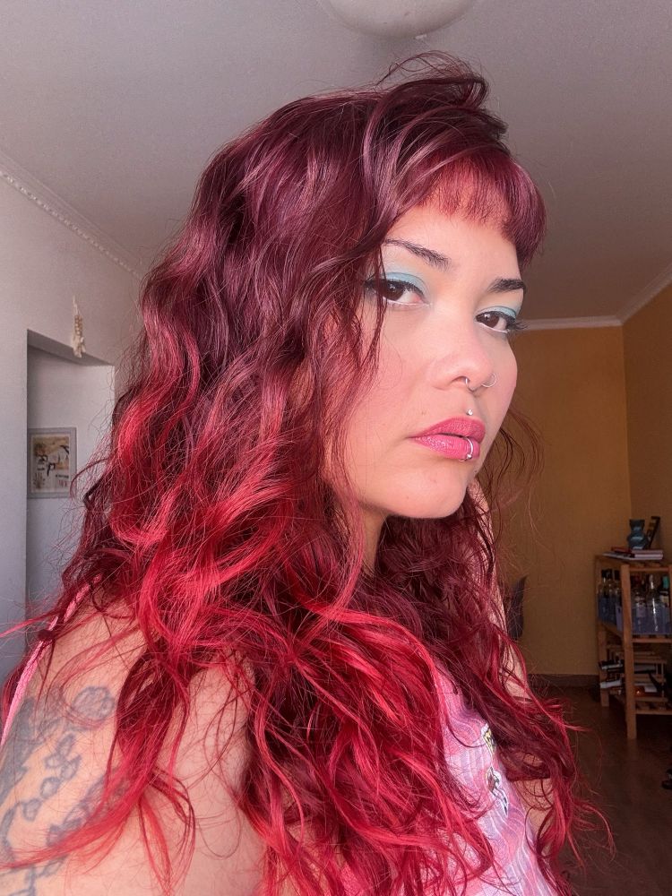 foto minha virada de lado olhando pra camera. sou branca, cabelos vermelhos, estou usando uma blusa rosa e tenho piercings visíveis no nariz e labio.