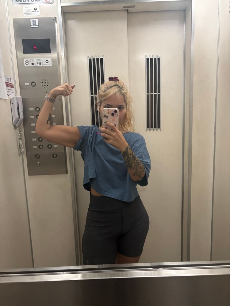 foto minha no espelho do elevador flexionando os músculos do bíceps. meu cabelo é loiro, estou vestindo uma blusa azul e um short cinza 
