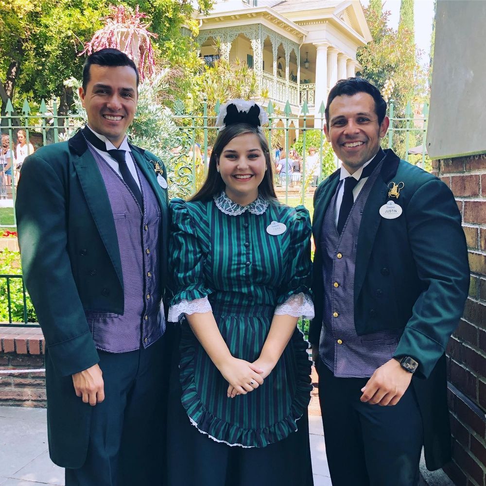 Three "cast members" (employees) who work at Disneyland's The Haunted Mansion in costume. They consist of either a dark green butler-like suit with a light purple vest, or a dark green striped dress with apron and headdress (resembling a simplified maid outfit)