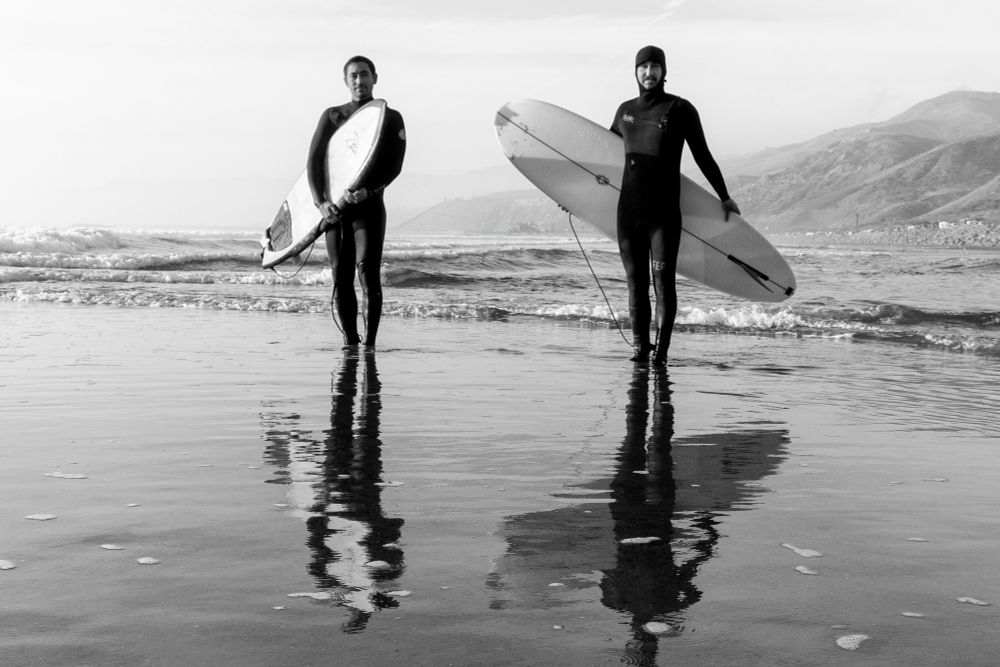 Two surfers standing in ankle shallow water showing their reflection below. Mountains in the background