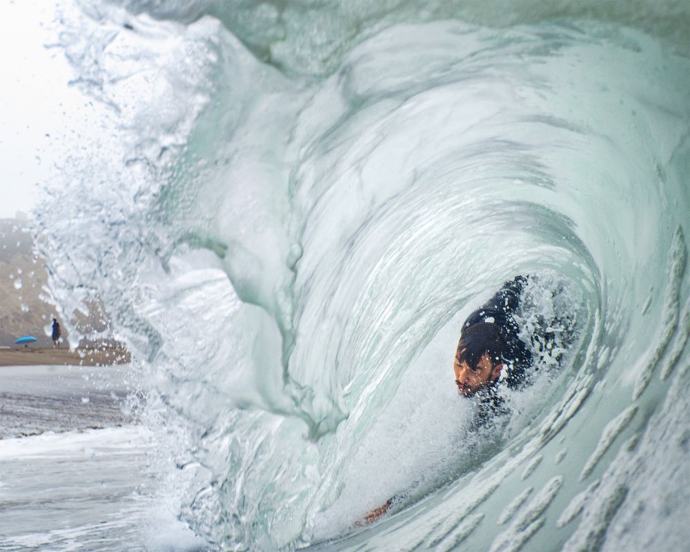 Bodysurfer in a barrel shore break. Person and blue umbrella seen on the beach just in the background