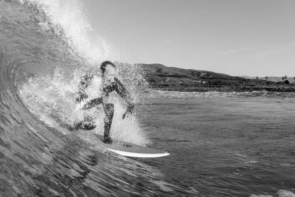 Surfer with some of the wave crashing over his back as he shoots down the wave. Coast in the background. Black and white photo