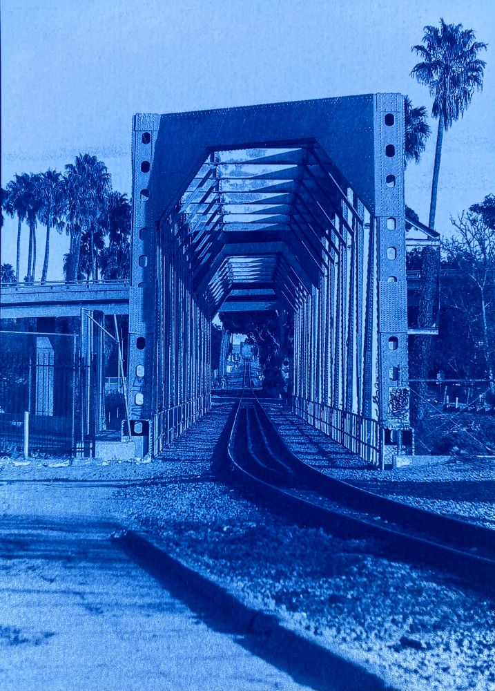 Cyanotype photo of looking down the train tracks off to the horizon through railroad bridge. Palm trees in the background