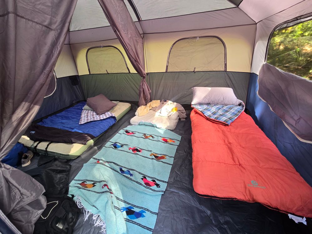 The interior of a 2-room tent.  There's a queen sized air mattress with a blue sleeping bag on it on the left, a cot with an orange sleeping bag on it on the right, and a dog bed and Mexican blanket in the middle.