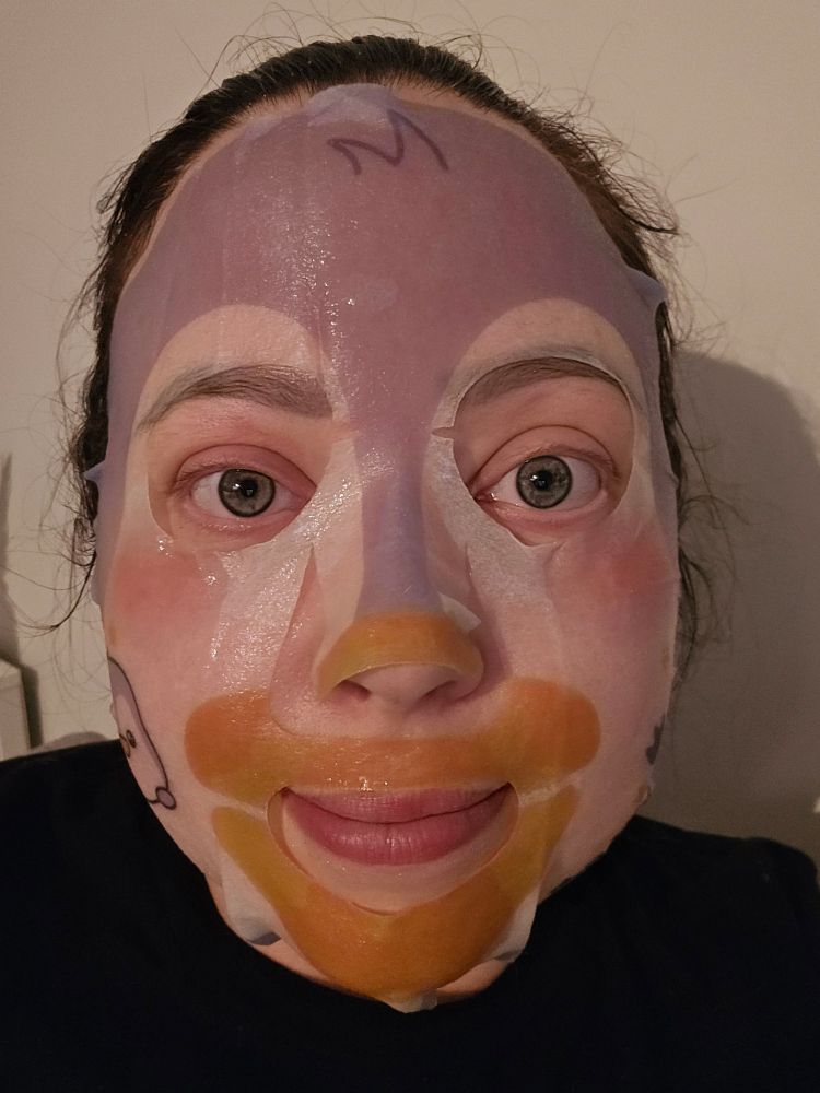 A brunette lady with no makeup on is wearing a printed sheet mask that is slightly resembling a penguins face, with an orange beak and lavender feathers and rosy little cheeks. She is smiling a little. 