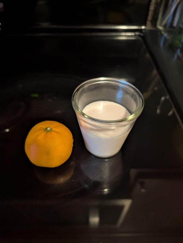 A small glass cup with a white liquid, next to a mandarin orange, on a black background.
