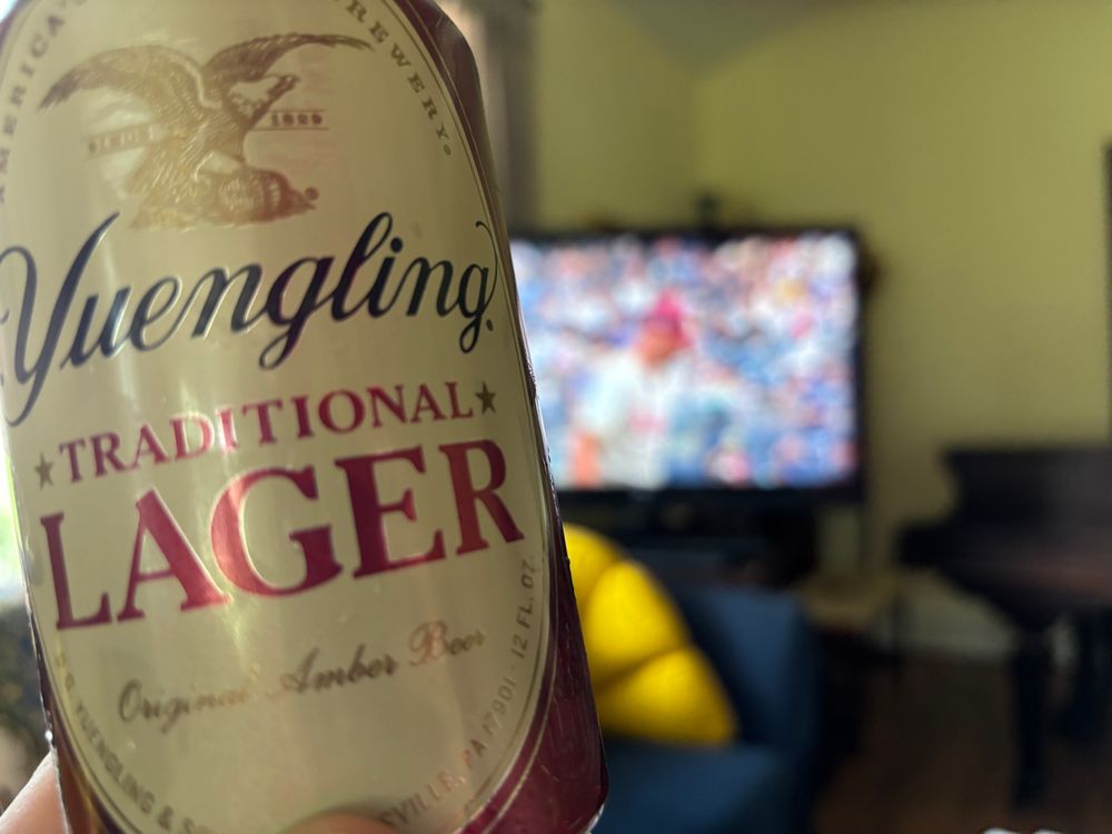Yuengling lager can being held in front of TV with blurry image of Phillies player on screen. 