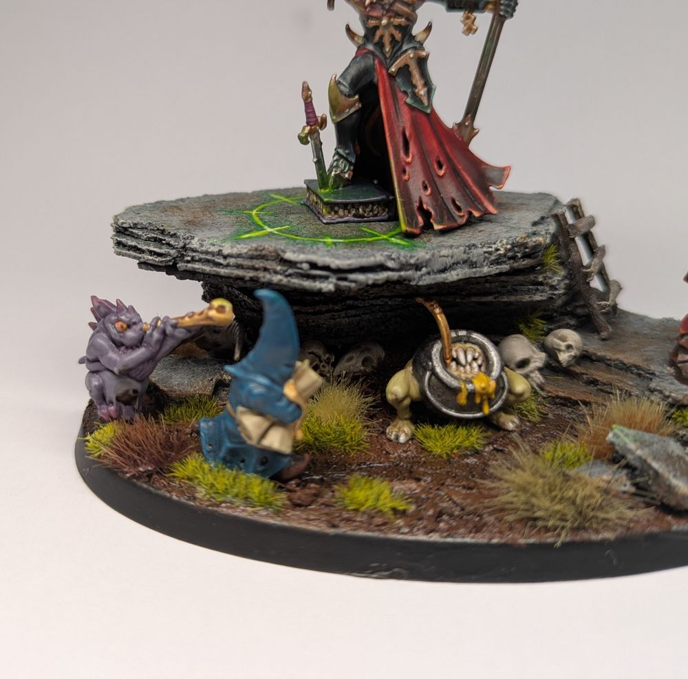 A purple lizard minion toots a horn in the ear of a blue wizard who clutches some scrolls. Behind them lurks a hungry toothed cauldron.