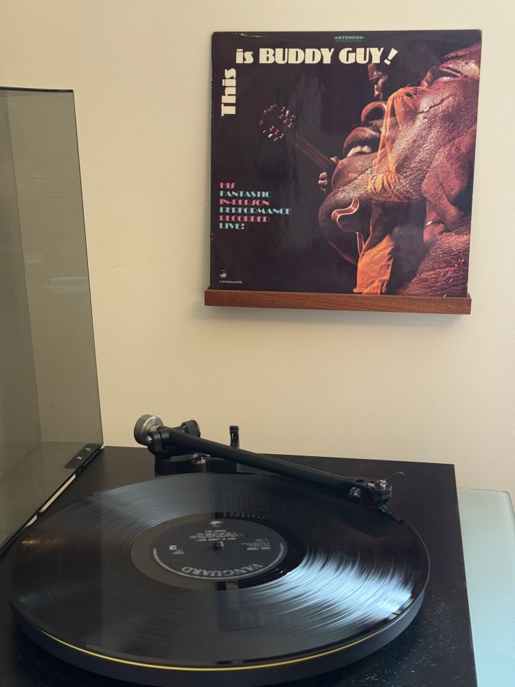 A black vinyl record spins on a turntable beneath the cover of This is Buddy Guy!, a live album by the legendary blues guitarist. The cover shows a dynamic photo of Buddy Guy performing, with bold text announcing “His fantastic in-person performance recorded live!” The setup highlights raw blues energy captured in an analogue format.