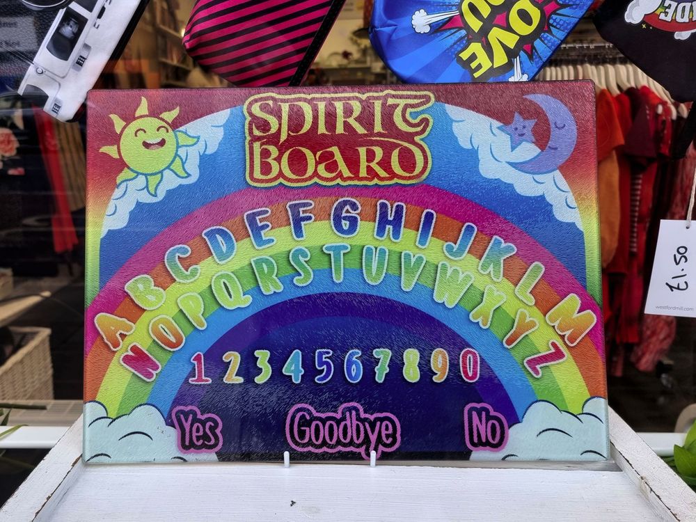 A ouija board displayed in a charity shop window. It features the usual alphabet, numbers and yes/no/goodbye keywords. However its styling is unusual. The font is cheerful and multi-coloured, the background is a rainbow, and the corners feature a happy sun and moon. The overall impression is of a toy intended for very young children.