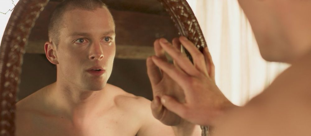 A white man with buzz cut hair touches his own reflection in a large oval mirror