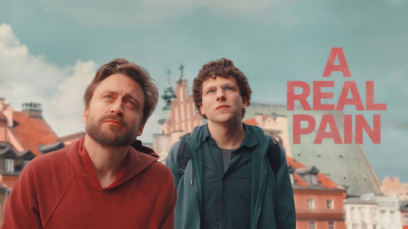 A photo of Kieran Culkin and Jesse Eisenberg as cousins on a trip to Poland, in Eisenberg's film, A Real Pain. The men are looking above the camera at a building in Warsaw. The name of the film is printed in pale red capitals to the right of the still.