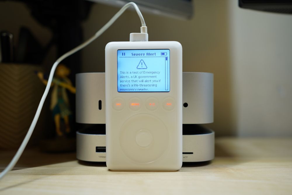 A 2003 iPod 3rd generation with a warning displayed on the screen. Title: "Severe Alert". Message: "This is a test of Emergency Alerts, A UK government service that will alert you if there's a life-threatening emergency nearby."