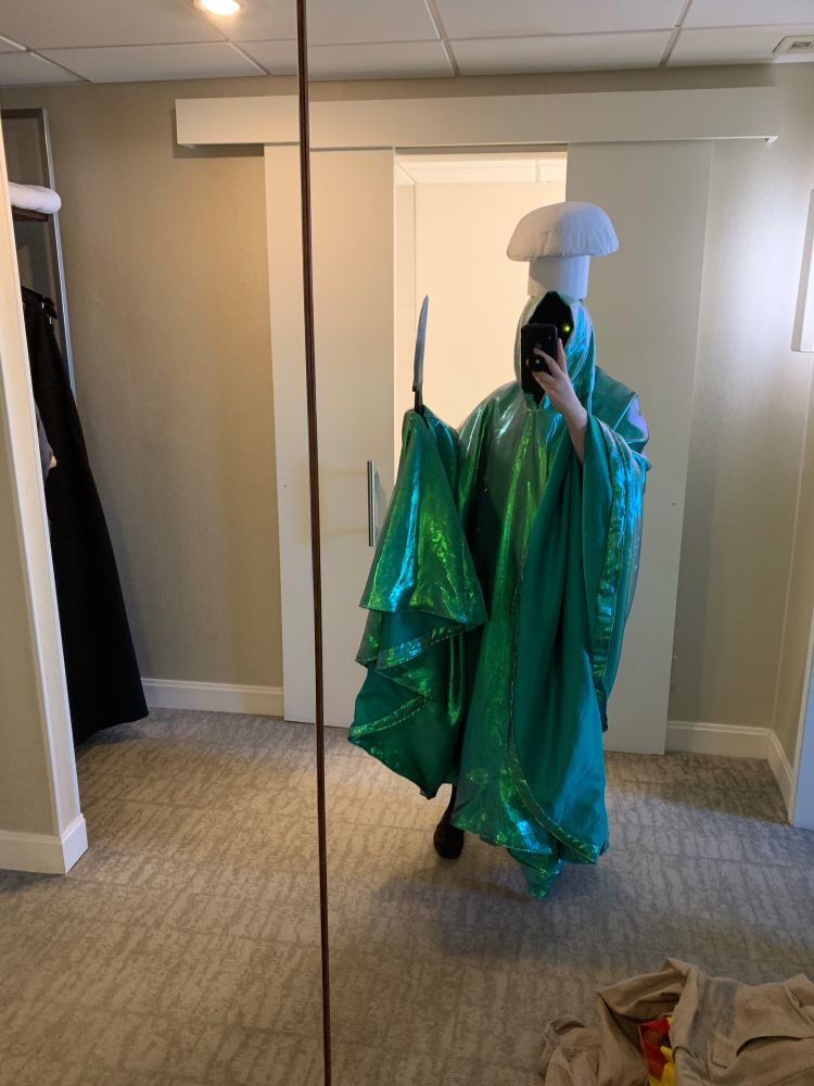 Sam wearing a full face mask of black with a long shiny green cloak holding a fake knife with a big chef hat.