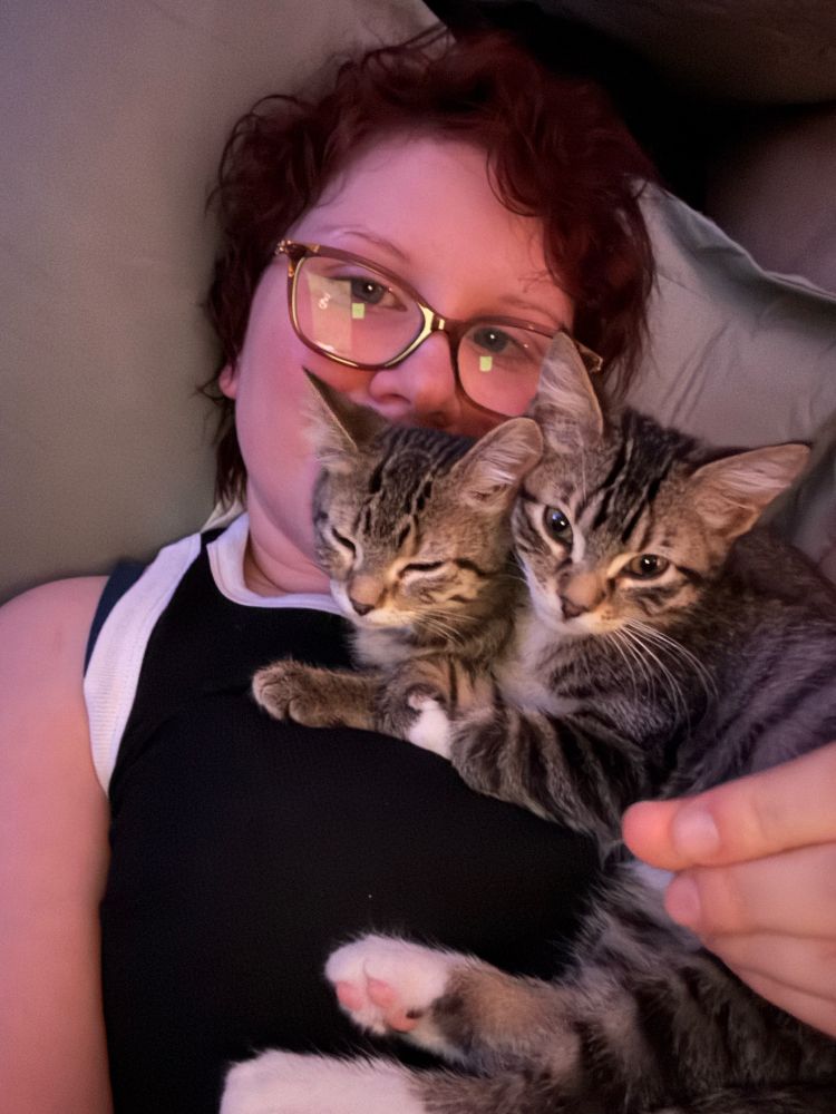 Two, tabby kittens laying in a pile on a woman with red hair, glasses, and a black tank top 