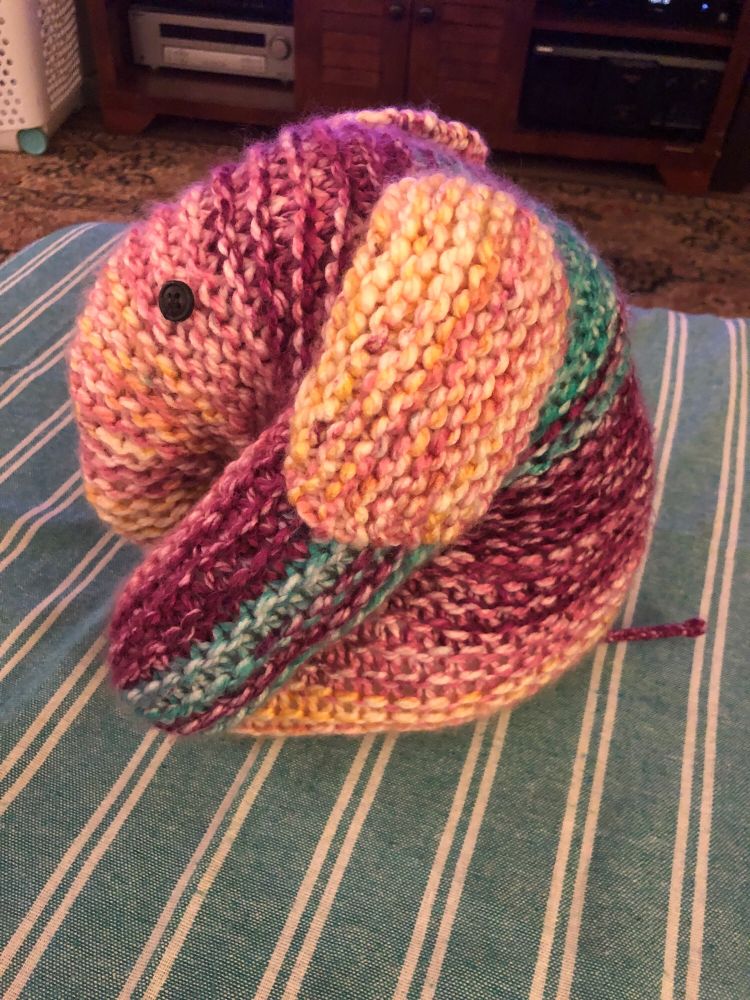 A knitted elephant plush sits on striped teal and white blanket. The plush has black button eyes, and the yarn is striped in pink, magenta, yellow, and teal. The elephant is in profile.