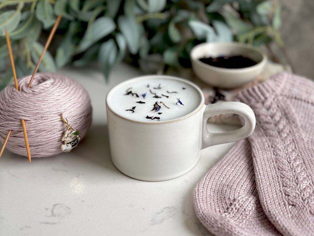 A small grey mug sits on a countertop, filled with a London Fog. Light purple cable knit socks and a matching cake of yarn sit on either side of it. 