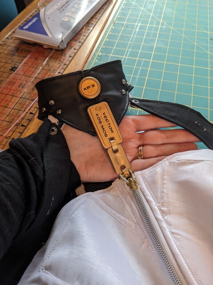 A photo of a hand holding up a zipper pull that has been attached to a collar on a bodysuit.  The pull is made of two links, with one labeled "VECTOR" and "KOS-MOS".  The collar also has a gold plaque that reads "KP-X".