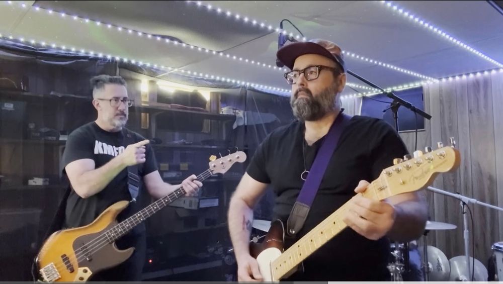 A bass player, wearing a KMFDM concert T, pointing at a guitar player steeling himself to play a wild solo.