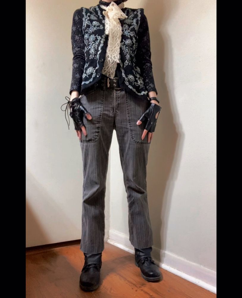 A photo of the same person cropping out the face. They are wearing a monochromatic outfit consisting of black boots, flared-leg cropped pinstripe charcoal trousers, a long sleeved black turtleneck with a pattern of white Christmas trees on it, a black wool vest with white embroidery, a bunch of white lace tied around the throat, and mismatched black fingerless gloves