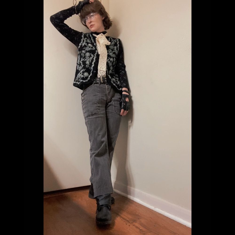 A photo of a young slim white person with Bob-length brown hair and glasses. They are wearing a monochromatic outfit consisting of black boots, flared-leg cropped pinstripe charcoal trousers, a long sleeved black turtleneck with a pattern of white Christmas trees on it, a black wool vest with white embroidery, a bunch of white lace tied around the throat, and mismatched black fingerless gloves