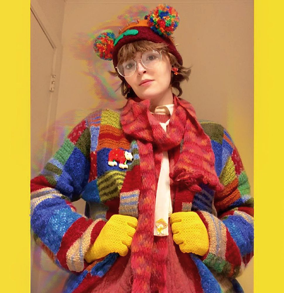 A photograph of a young slim white person with round glasses and bob-length brown hair. They are wearing an outfit which has a cozy-looking cardigan on multiple patchwork colors, bright yellow gloves, mushroom earrings, a burgundy knit hat, a coral knit scarf, a brooch of a polar bear wearing a clown suit, and a headband over the hat which has spikes and colorful pom-poms.
