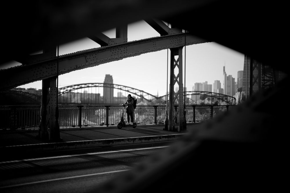 A young woman drifts across a bridge on the outskirts of Frankfurt on her e-scooter. Her silhouette is framed by the bridge structure. In the background is another bridge and the Frankfurt skyline.