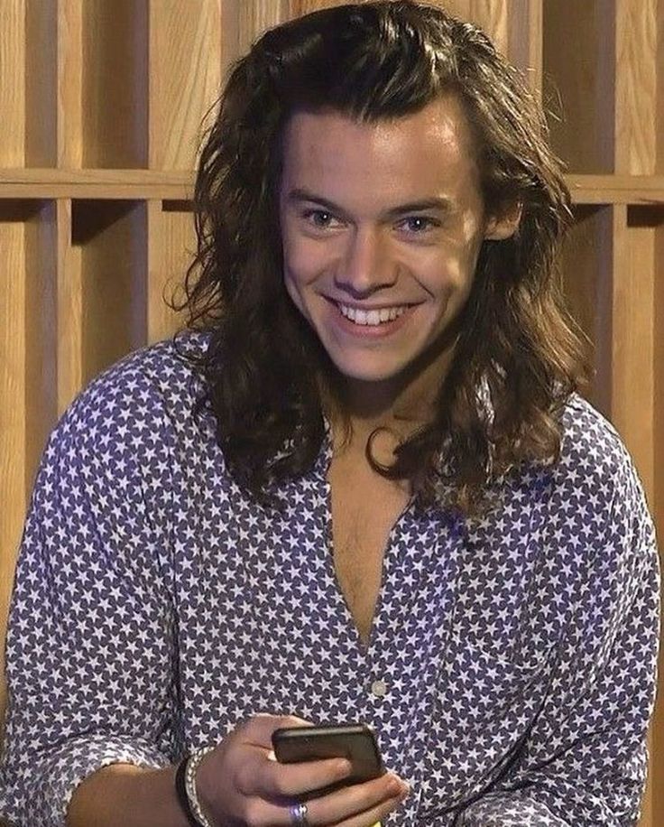 Harry Styles with long hair, wearing a blue button up with white stars, holding his phone, smiling 
