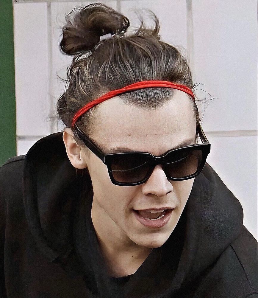 Harry Styles with his long hair in a bun, wearing a black hoodie, sunglasses and a red headband. 