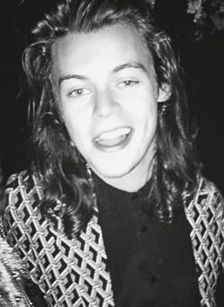 Close-up black and white picture of Harry Styles with long hair, smiling at the camera. 
