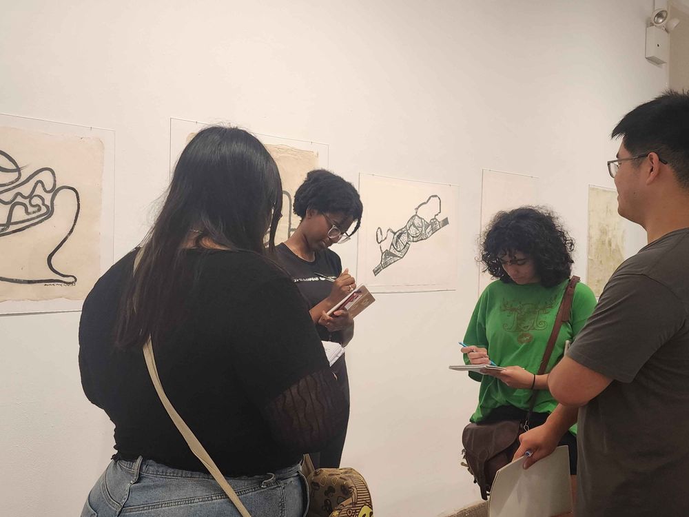 College students taking notes while in front of multiple black and white drawings pasted on the wall of a gallery.