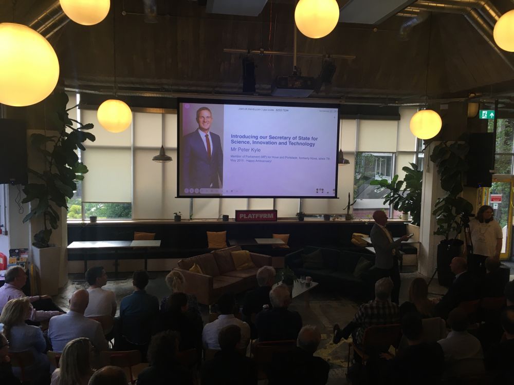 Tiered steps with a screen showing a picture of Peter Kyle with people waiting.