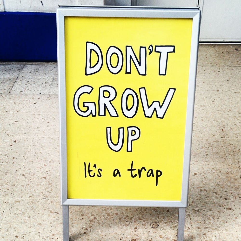 A yellow standup sign with handwritten like printed text that reads “Don’t grow up” and in smaller letters “it’s a trap”