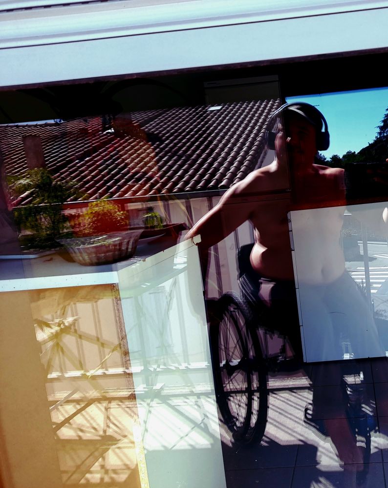 Selfie par reflet sur la vitre d'une porte-fenêtre. Un homme torse nu assis sur un fauteuil roulant. La lumière rasante accentue la définition des muscles de ses épaules et bras. 