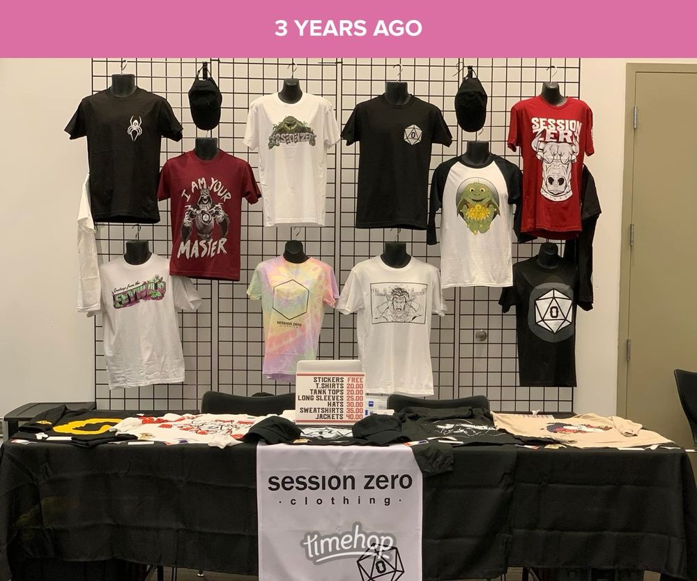 A convention booth with several articles of clothing hanging on wire racks and laying on a table. There is a black tablecloth and a white table runner laying on top of it with “Session Zero Clothing” written in large font.