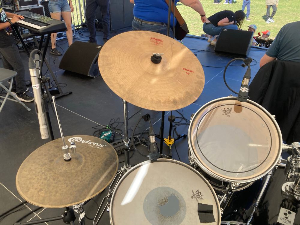 Des futs et cymbales d'une batterie 🥁 sur scène, depuis la place du batteur. Y'a des micros 🎤 et des câbles qui courent par terre, on aperçoit aussi un clavier 🎹.