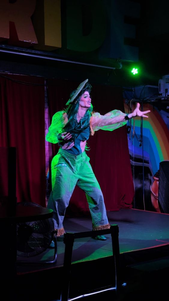 A drag performer onstage reaching out with one arm, dressed in green with Medicare branded pants