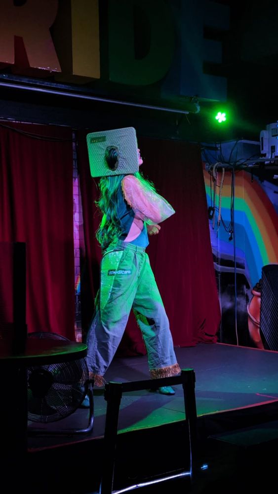 A drag performer onstage, you can't see their face because there's a giant Medicare card on their head (like a green credit card)