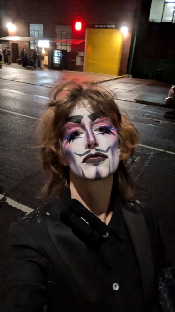 A selfie of a drag king in dark clothes on a damp street at night