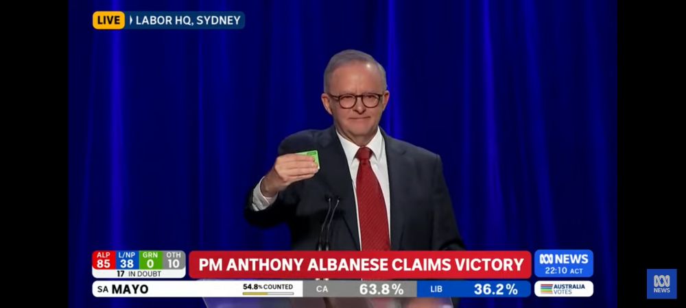 A screenshot of Australian election coverage with the words PM ANTHONY ALBANESE CLAIMS VICTORY and a middle aged man with glasses and a suit holding a green card
