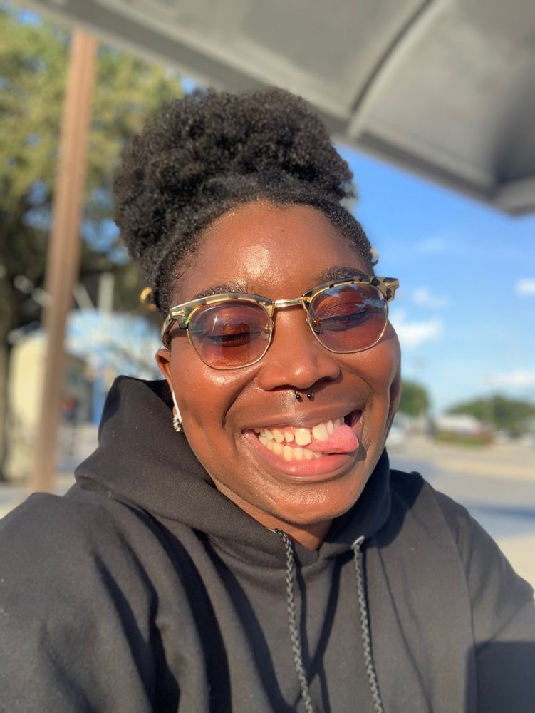 A person with dark skin, curly hair in a high puff, and tortoiseshell sunglasses smiles playfully while sticking out their tongue. They are wearing a black hoodie, and the sun casts a warm glow on their face. The background shows a clear blue sky and a street scene slightly out of focus.