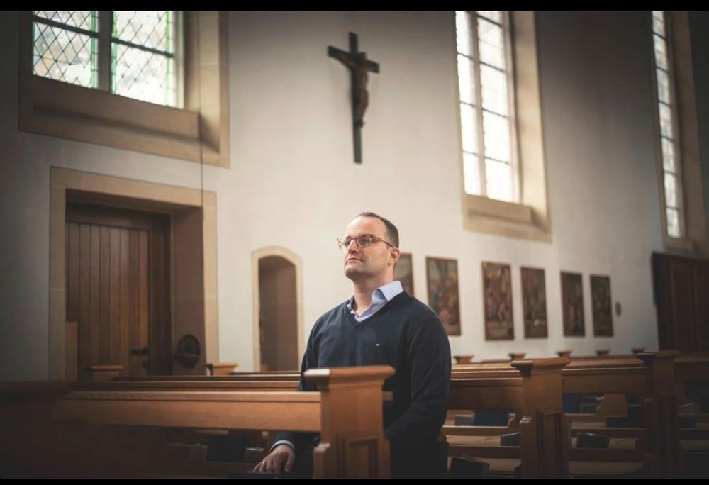 Spahn in einer Kirche, in der Bank, gerade Rücken,  trotziger Blick mit hoch gerecktem Kinn, Kruzifix über ihm. 